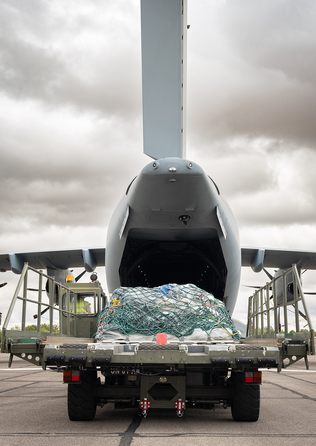 The Condec loader, holding the net-wrapped cargo pallet, aligns with the open rear cargo bay of an Atlas (A400M) aircraft, under a cloudy sky.